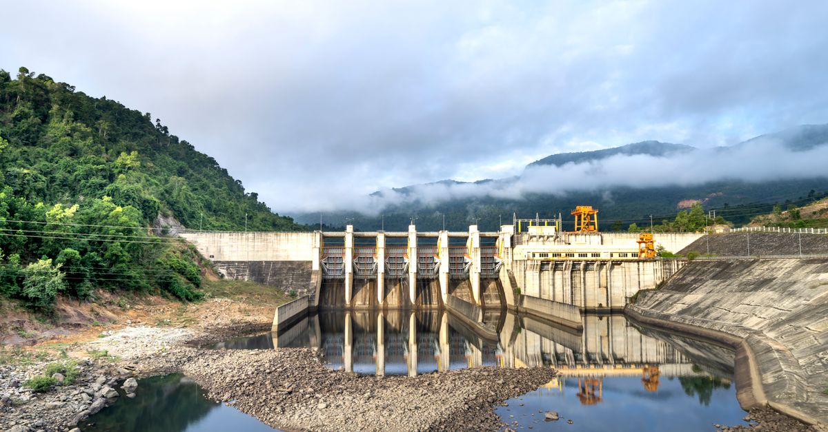 Industrial hydroelectric power plant located near river and stony coast against green mountain range covered with mist on summer day